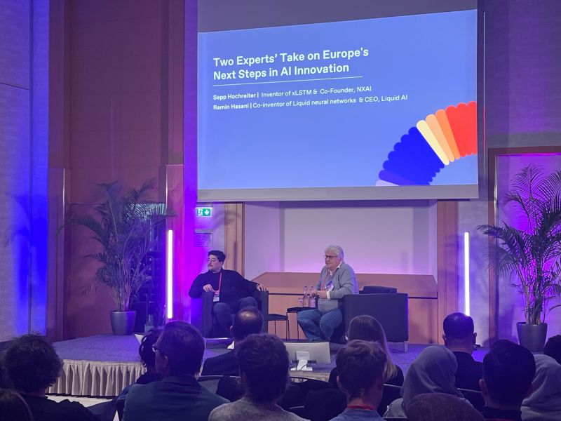 Two conference speakers sitting on stage, participating in a dialogue while attendees observe. Left in picture Ramin Hasani, Founder of Liquid AI, next to him on stage (right in picture) is Sepp Hochreiter, Founder of NXAI.